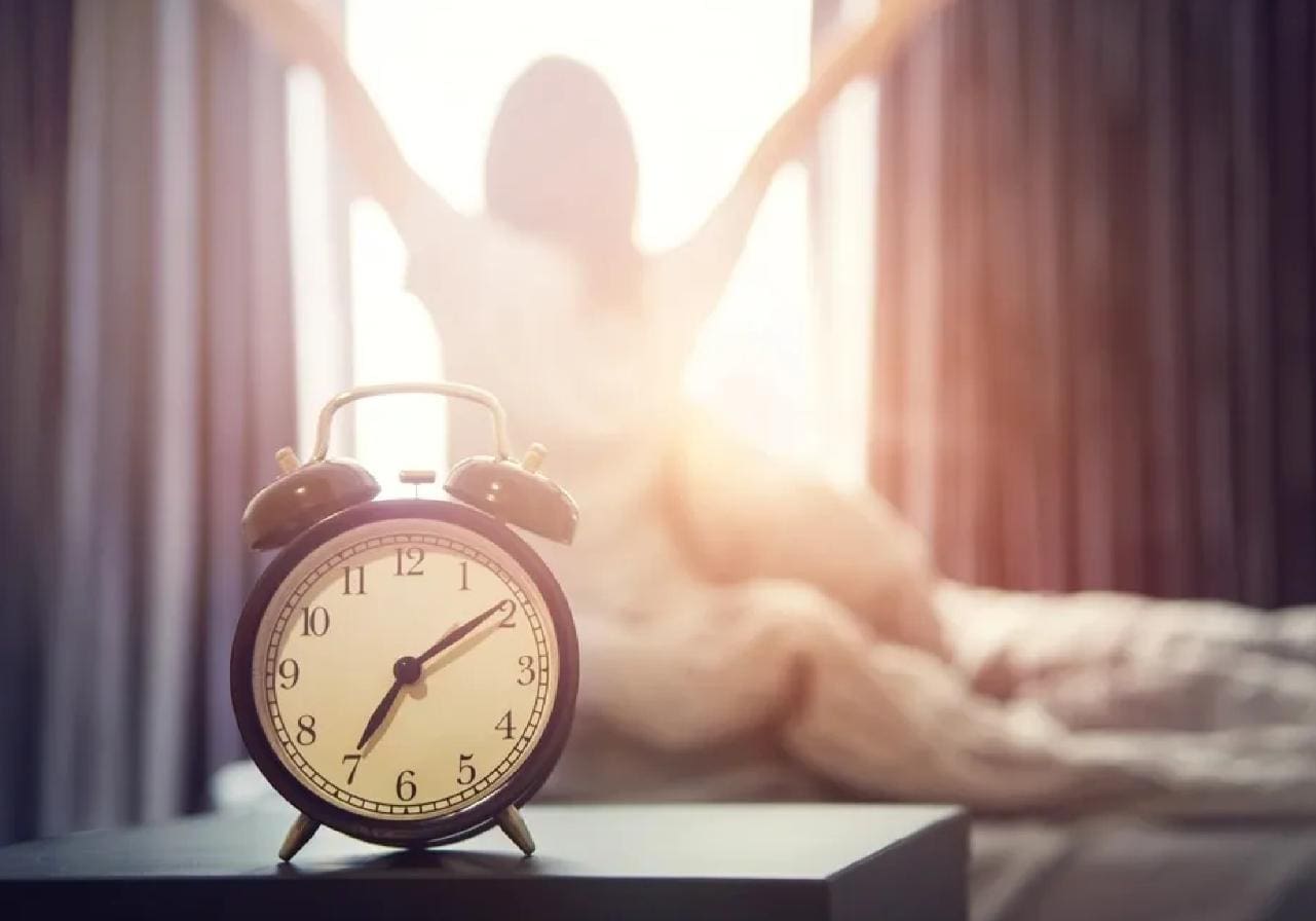 A woman is sitting in bed with her arms raised behind the alarm clock.
